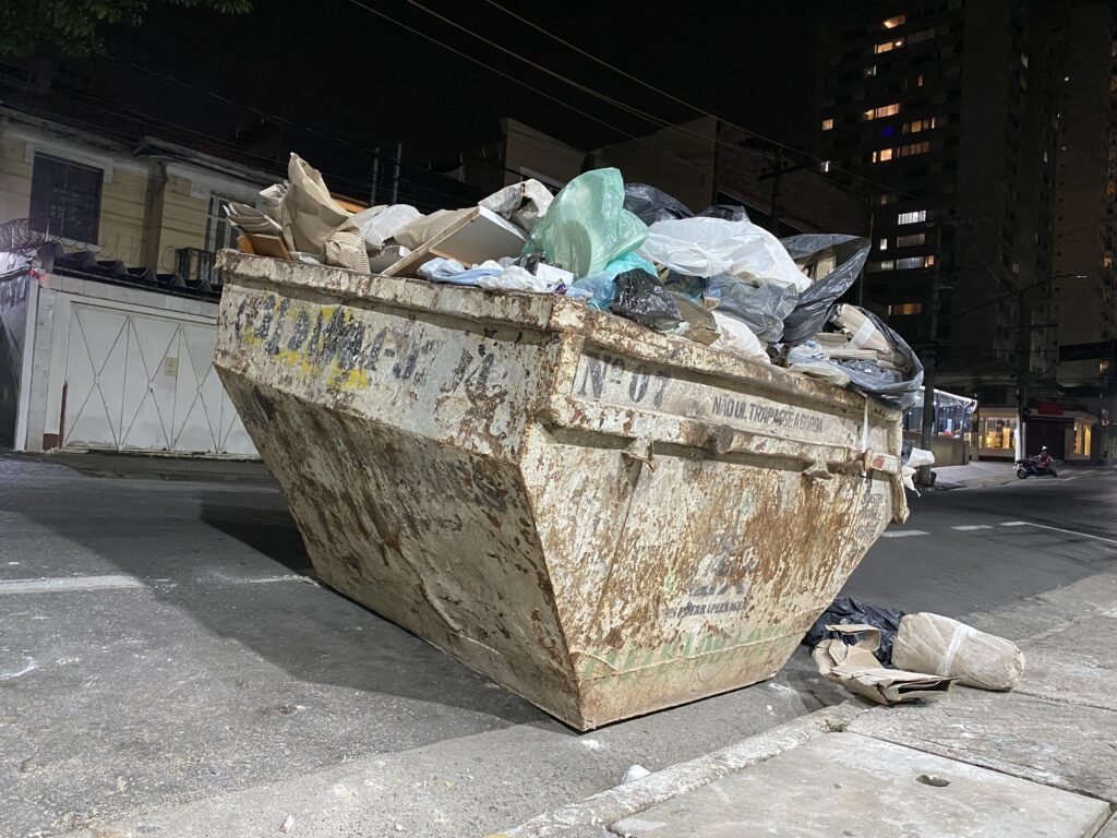 Caçamba estacionária com muito lixo e resíduos acima da borda